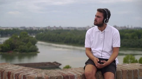Young man sits on brick wall and enjoy in music from cellphone, tugboat on river Stock-Footage 83689598