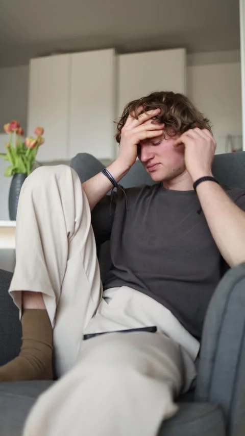 A young man sits in a chair, looking up while holding a phone. He appears frustr Stock Footage 302632601