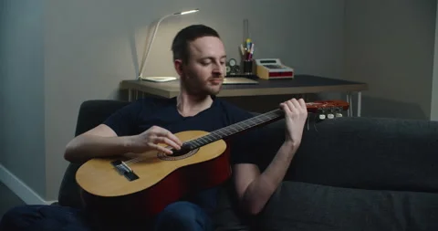 A young man sits on the couch and plays the guitar. Close-up portrait of a man Stock Footage 148133426