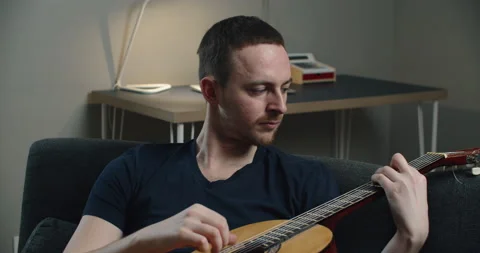 A young man sits on the couch and plays the guitar. Close-up portrait of a man Stock Footage 148133736