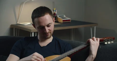 A young man sits on the couch and plays the guitar. Close-up portrait of a man Stock Footage 148292234
