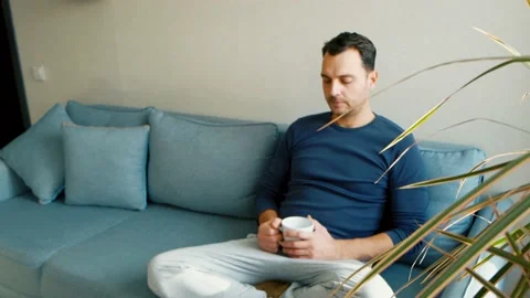 A young man sits on the couch holding a cup in his hands and drinking from it Stock-Footage 169302864