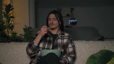 A young man sits on the couch at home and eats chips, actively gesturing Vidéo 170423583