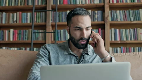 Young Man Sits On The Couch With Laptop And Talks On Cell Phone. Stock Footage 140224924