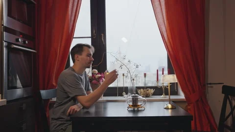 A young man sits down at a table by the window to drink tea Stock Footage 295171541