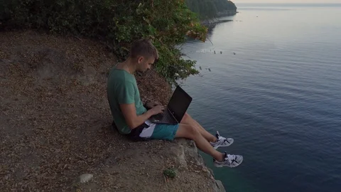 A young man sits on the edge of a cliff and works on a laptop. A man sits with a Video stock 117634890
