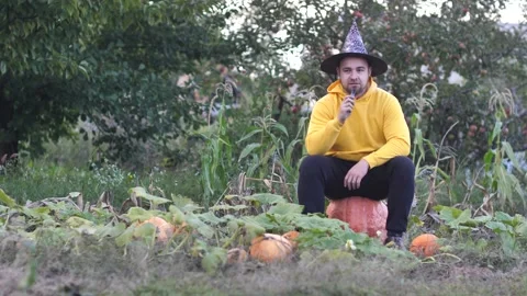 A young man sits on a huge pumpkin in a pumpkin field in a witch's hat and lets Stock Footage 213057288