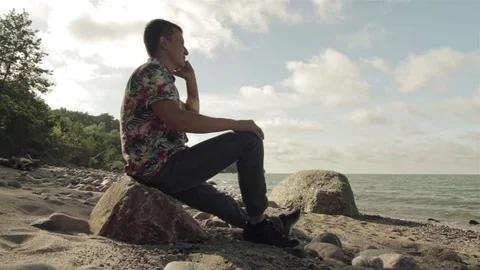 A young man sits on a large rock and talks on the phone by the sea. Stock Footage 78707101