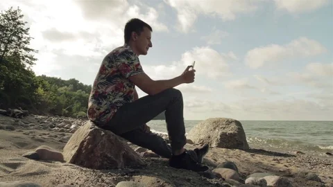 A young man sits on a large stone and looks at a mobile phone by the sea. 스톡 동영상 78705744