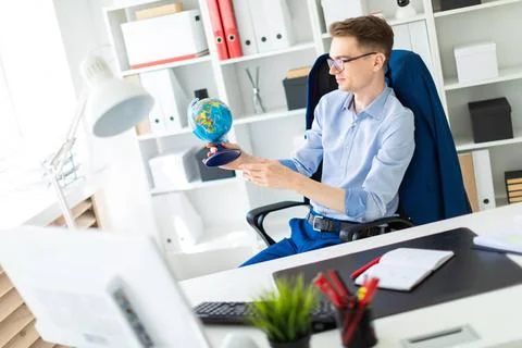 A young man sits in the office at a computer desk and holds a globe in his... Stock-Fotos