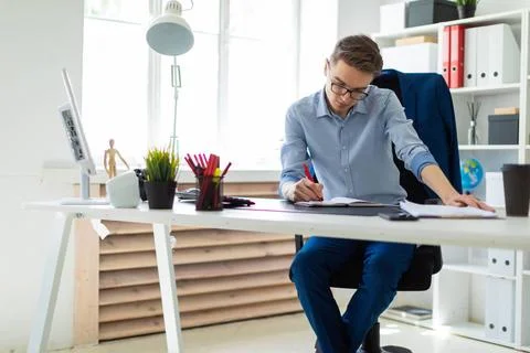 A young man sits in the office at a computer desk and writes in a notebook... Stock-Fotos