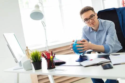 A young man sits in the office at a computer desk and holds a globe in his... Stock-Fotos