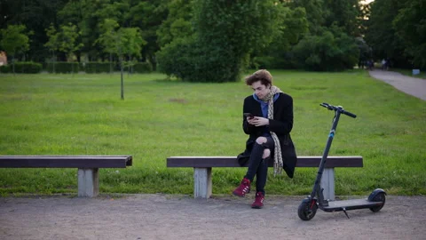 A young man sits on a park bench, gets up, leaves on an electric scooter. Vidéo 201076331