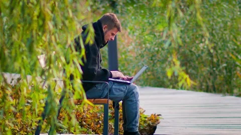 A young man sits on a park bench with a laptop on his lap, typing intently Stock Footage 287754674