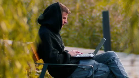 A young man sits on a park bench with a laptop on his lap, typing intently Stock Footage 287754697