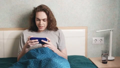 A young man sits reclining in bed and plays something on a mobile phone Stockbeeldmateriaal 196812443