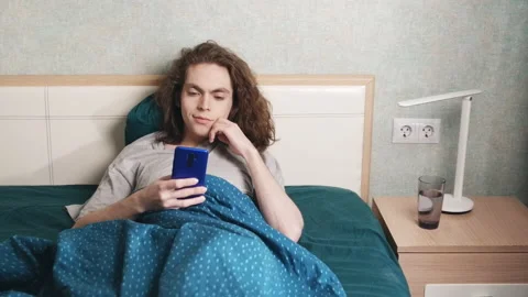 A young man sits reclining in bed and looks at the phone, then smiles a little 库存影片 196812533