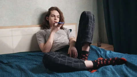 A young man sits reclining in bed and records a voice message for someone. 動画素材 196813046