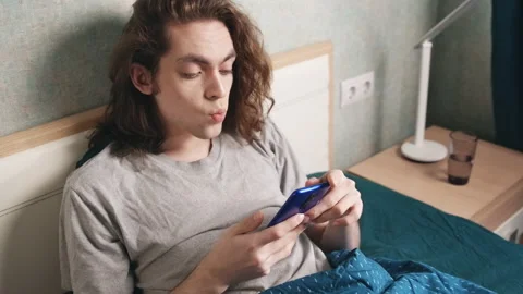 A young man sits reclining in bed and looks at the screen of the phone 库存影片 196814049