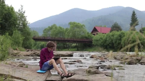 Young man sits on a rock in the river and draws of landscape Stock Footage 68940433