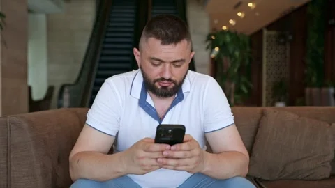 A young man sits on a sofa and holds a phone in his hands 動画素材 334376830