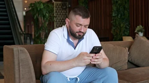 A young man sits on a sofa and holds a phone in his hands Stock Footage 334376962