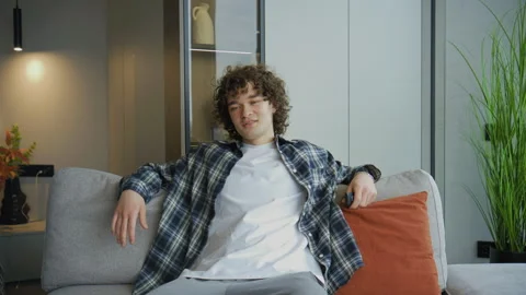 Young man sits on sofa and watches TV while holding remote control in modern liv Stock Footage 345297861