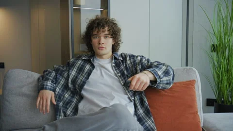 Young man sits on sofa and watches TV. He holds a remote control while watching  Stock Footage 345299122