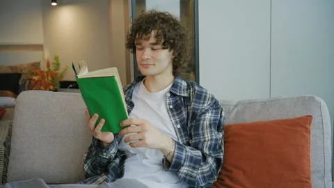 Young man sits on sofa and reads book in modern living room while enjoying free  Stock Footage 345300804