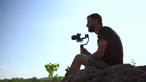 A young man sits on a stone with a camera in his hands. Man shoots at camera Stock Footage 136439890