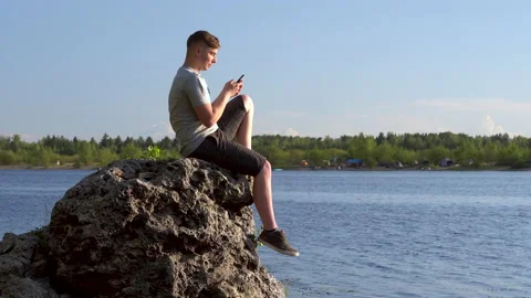 A young man sits on a stone by the river with a phone in his hands. The man is Stock Footage 136438728