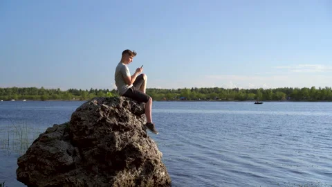 A young man sits on a stone by the river with a phone in his hands. The man is Stock Footage 136438989