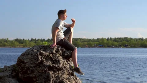 A young man sits on a stone by the river and admires nature. A man in nature Stock Footage 136439064
