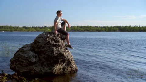 A young man sits on a stone by the river and admires nature. A man in nature. Stock Footage 136439637