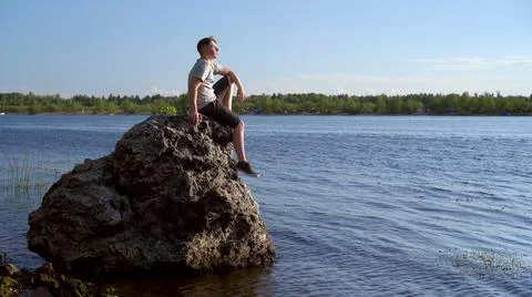 A young man sits on a stone by the river and admires nature. A man in nature. Stock Photos