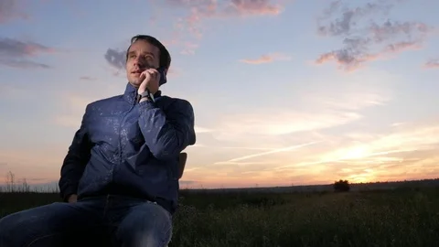 A young man sits at sunset and talks on his mobile phone. Beautiful sky behind. Stock Footage 76922525