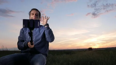 A young man sits at sunset and does selfie on a mobile phone. Beautiful sky Stock Footage 76922700