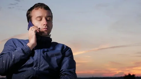 A young man sits at sunset and talks on his mobile phone. Beautiful sky behind. Stock Footage 76923151