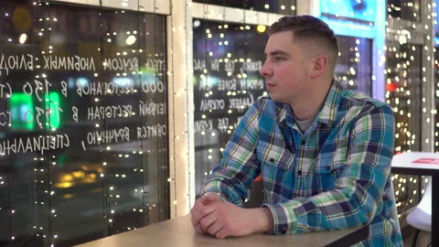 A young man sits at a table in a cafe and looks out the window. The guy sits in Stock Footage 166677892