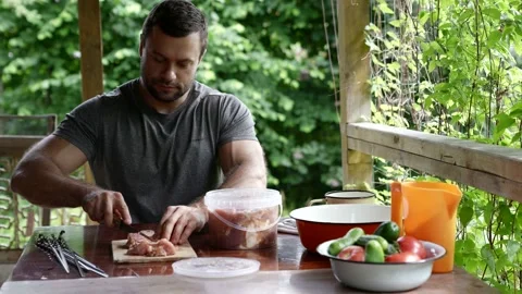 A young man sits at a table in a gazebo and cuts raw meat into pieces Stock Footage 163661037