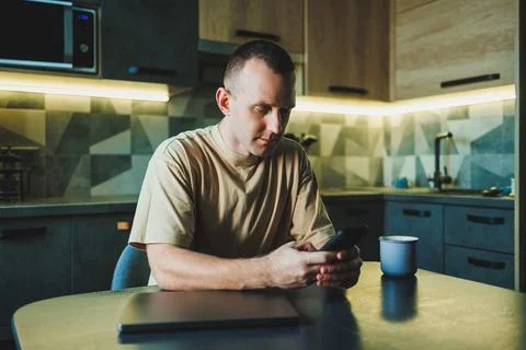 A young man sits at the table in the kitchen and talks on the phone. The lapt Stock Photos