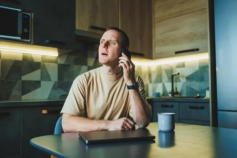 A young man sits at the table in the kitchen and talks on the phone. The la.. Stock Photos