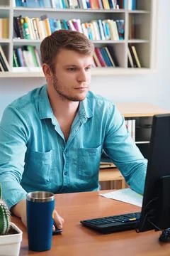 Young man sits at table in room with books and looks into computer monitor Foto stock