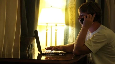 A young man sits at a table. Works at the computer. And talking on a cell phone. Stock Footage 97084024