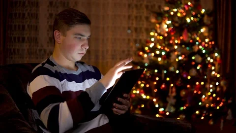 A young man sits with a tablet in him hands on the background of a Christmas Stockbeeldmateriaal 121232474