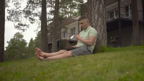 A young man sits under a tree and works on a laptop. A man with a laptop in Stock Footage 246048271