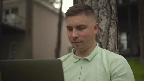 A young man sits under a tree and works on a laptop. A man with a laptop in Stock-Footage 246048279