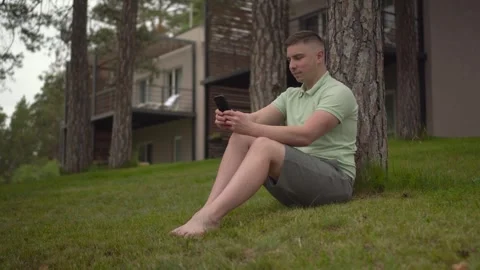 A young man sits under a tree near a country house with a phone in his hands. A Stock Footage 246356679