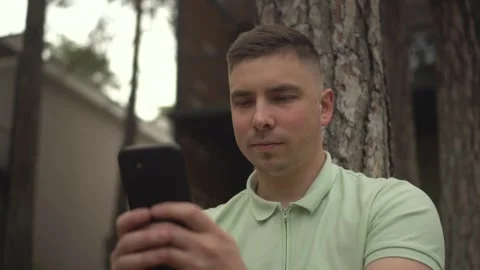 A young man sits under a tree near a country house with a phone in his hands. A Vídeos de archivo 247628784