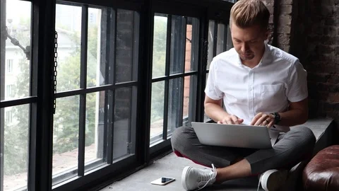 A young man sits on a windowsill and works using a laptop and a smartphone. Stock Footage 97045127